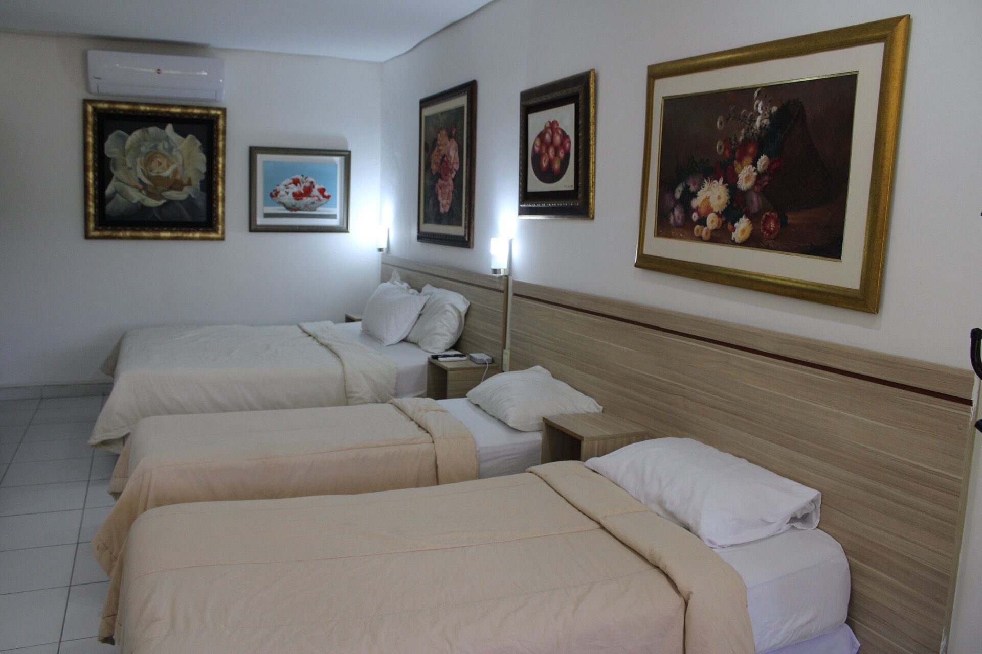 Una habitación de hotel con tres camas individuales, una mesita de noche con lámpara y cuadros en las paredes.