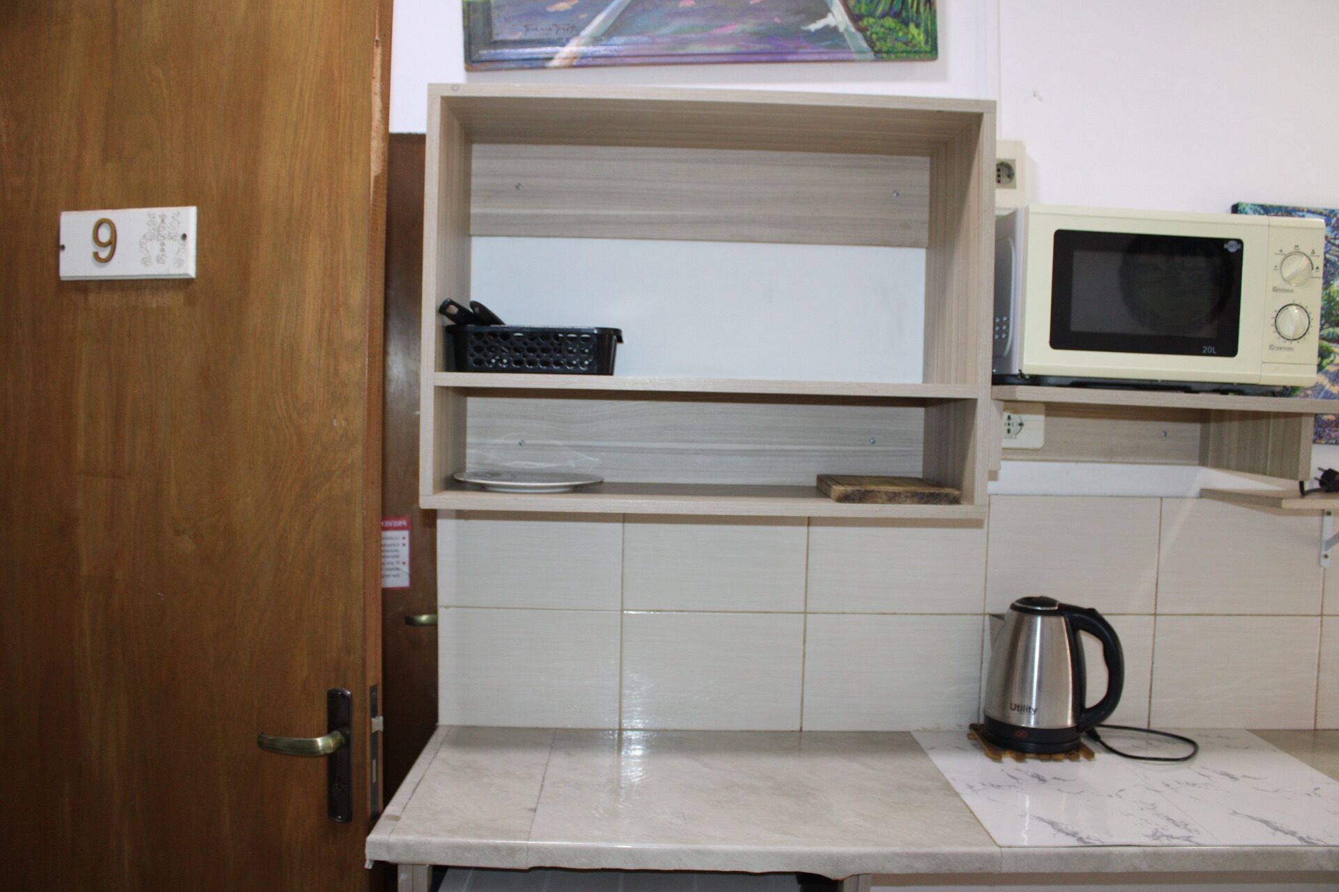Una cocina con puerta de madera, microondas, hervidor y una estantería.