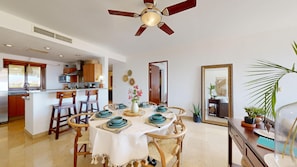 A dining area with a round table set for four, surrounded by wooden chairs. A ceiling fan is visible above, and the room has a kitchen in the background.