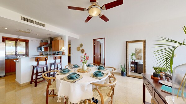 A dining area with a round table set for four, surrounded by wooden chairs. A ceiling fan is visible above, and the room has a kitchen in the background.