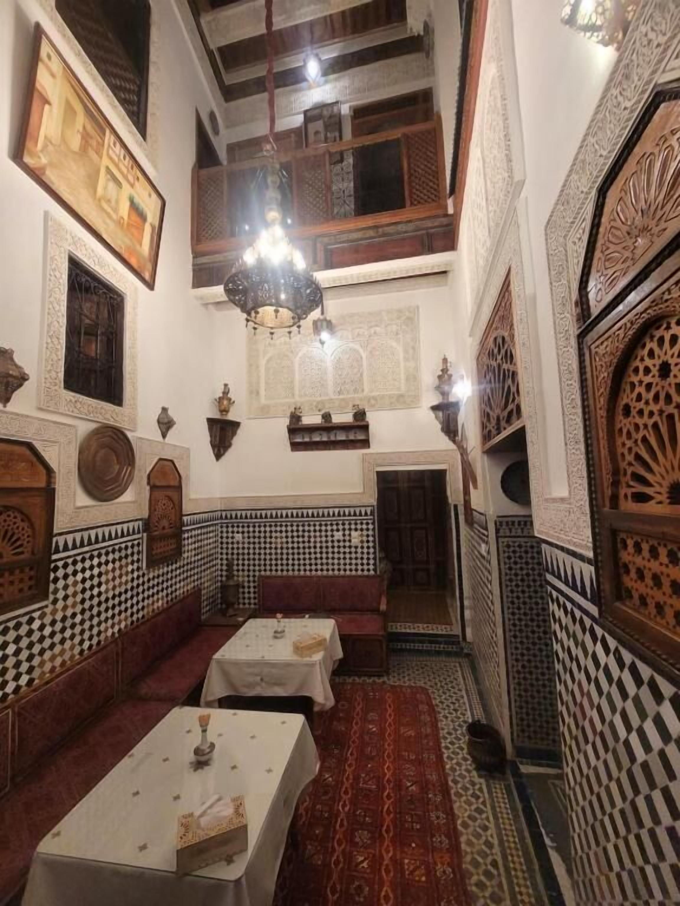 Um corredor estreito e ricamente decorado, com paredes de azulejos xadrez, tapete estampado e uma série de mesas e bancos.