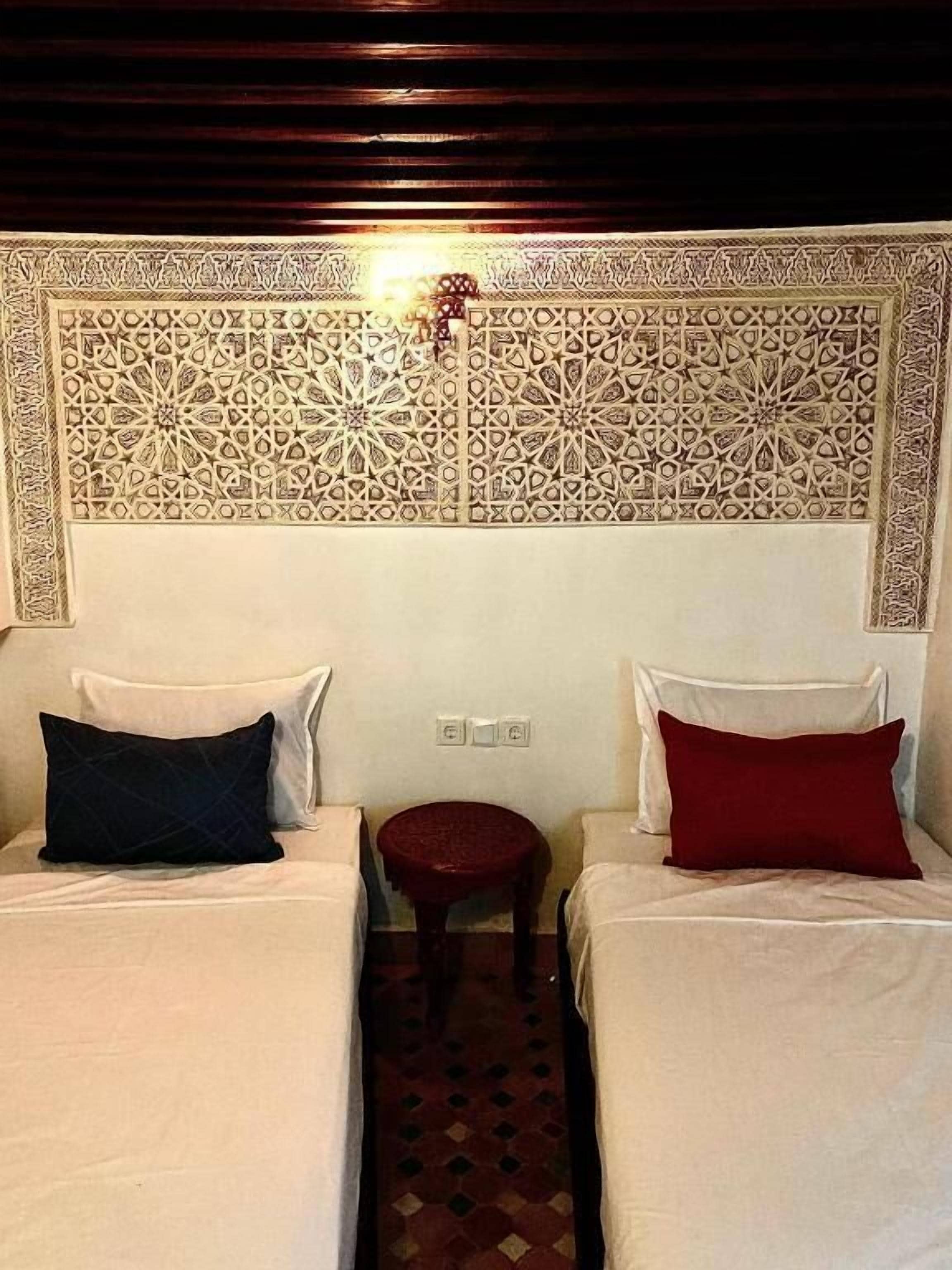 A room with two beds, each with a pillow, a small round table, and a decorative wall panel.
