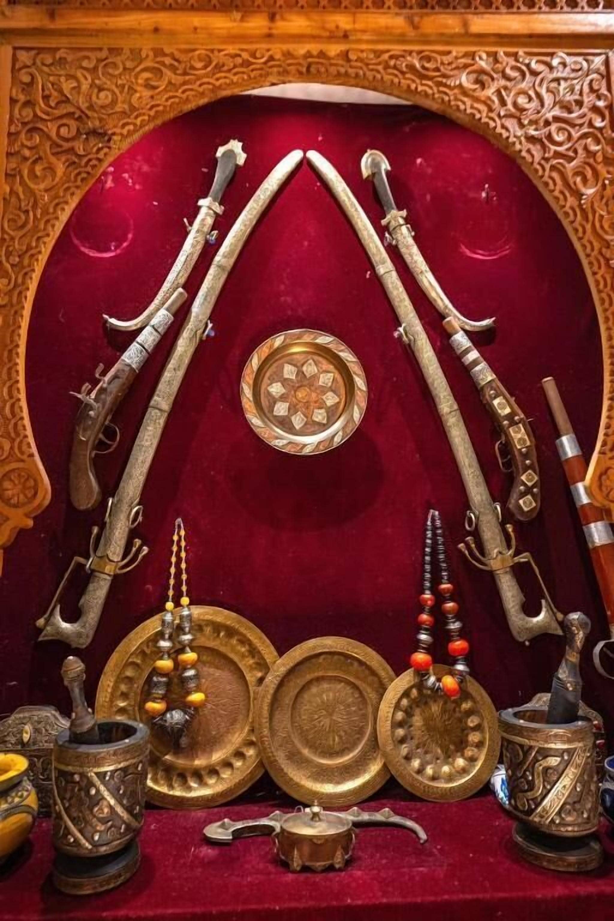 Une exposition d’armes anciennes et d’assiettes décoratives, présentées sur un fond de velours rouge agrémenté de sculptures en bois ouvragées.