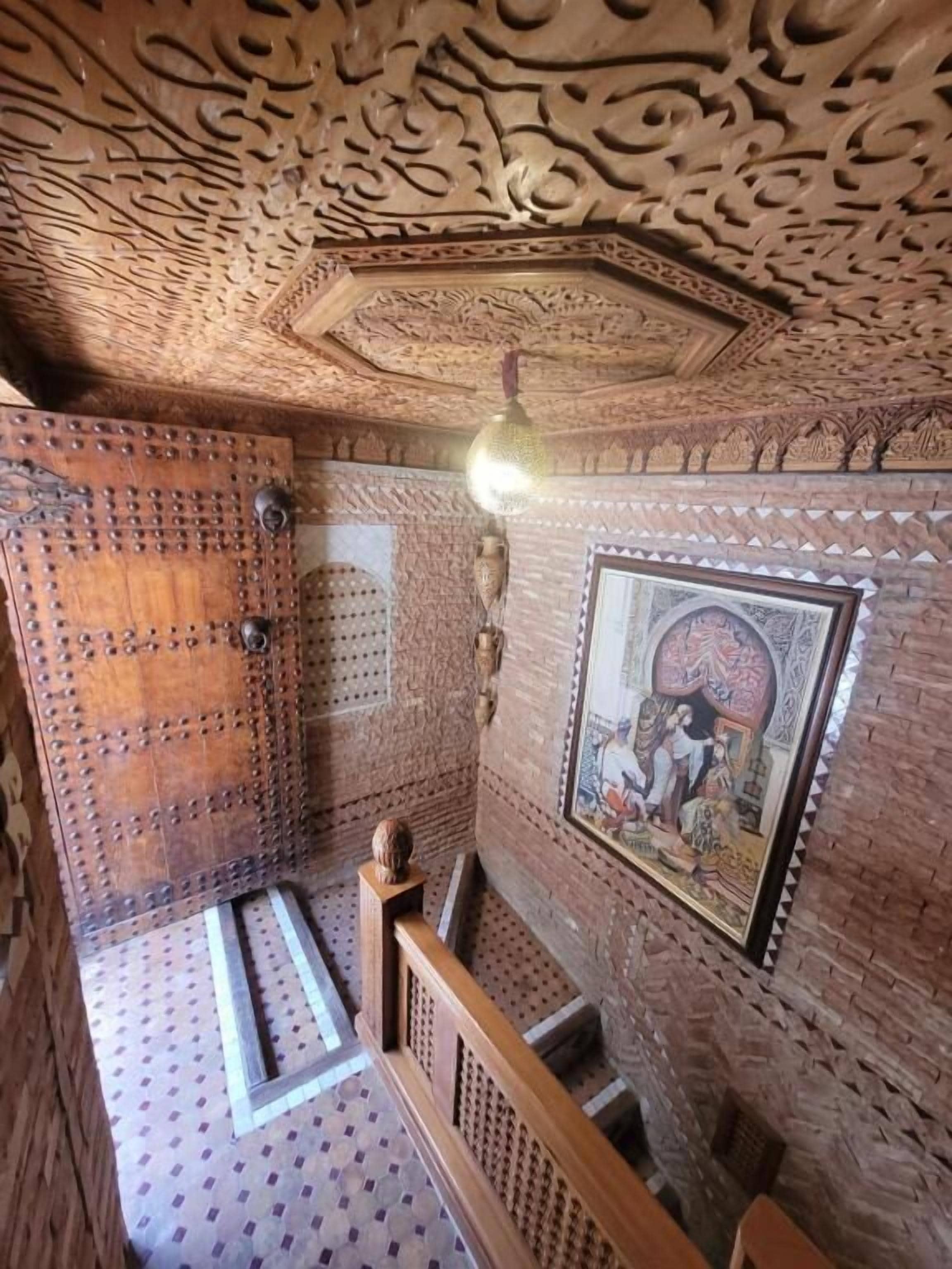 Een kamer met gedetailleerde houtsnijwerken aan het plafond en de muren, een houten deur met metalen knopen, een schilderij in een lijst en een vloer met een patroon.