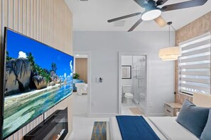 Una habitación de hotel moderna con una televisión grande, una cama, un ventilador de techo y un baño con cabina de ducha de vidrio.