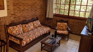 A room with a brick wall, a wooden sofa with patterned cushions, a wooden armchair, a small table with a laptop and a drink, and a window with a view of trees. - Vacances Centro (Pinamar)