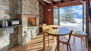 Une pièce chaleureuse avec un mur en pierre, des poutres en bois, une table ronde en bois et une vue sur des montagnes enneigées.