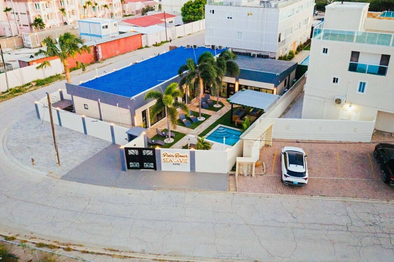 A hotel with a swimming pool, palm trees, and a sign for 'Palm Beach Sea Lavie Suites'.