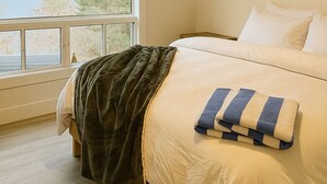 Ein Schlafzimmer mit großem Fenster, einem Bett mit weißer Bettwäsche, einer dunklen Tagesdecke und Blick auf einen See und Bäume.