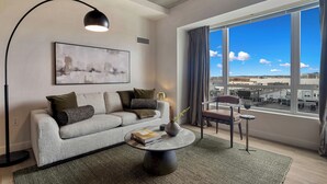 A modern living room with a grey sofa, a round coffee table, and a floor lamp. A large window offers a view of buildings and a clear sky. - Lux 2BR in Assembly Square | Pool & Fitness Center (Somerville)