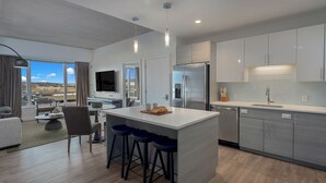A modern kitchen with a central island, bar stools, a large refrigerator, and a dining area with a view. - Lux 2BR in Assembly Square | Pool & Fitness Center (Somerville)