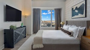 A modern hotel room with a large bed, a flat-screen TV, a console table with a plant, and a city view through the window. - Lux 2BR in Assembly Square | Pool & Fitness Center (Somerville)