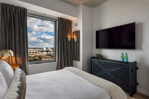 A hotel room with a large bed, a TV mounted on the wall, and a view of the cityscape through the window. - Stylish 1BR in Assembly Square | Pool, Gym & Views (Somerville)