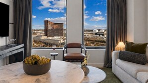 A modern hotel room with a large window, a dining table with a bowl of artichokes, a chair, a sofa, and a TV. - Stylish 1BR in Assembly Square | Pool, Gym & Views (Somerville)