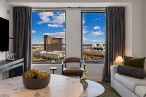 A modern hotel room with a large window, a dining table with a bowl of artichokes, a chair, a sofa, and a TV. - Stylish 1BR in Assembly Square | Pool, Gym & Views (Somerville)