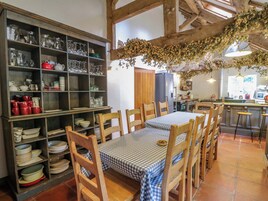 A dining area with a wooden table set for four, surrounded by chairs. A large wooden cabinet filled with dishes and glasses is to the left.
