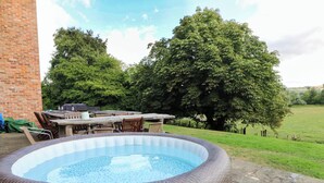 An outdoor hot tub with wicker walls, surrounded by a wooden deck, a brick wall, and a grassy field with trees. - Gaer Hall (Welshpool)
