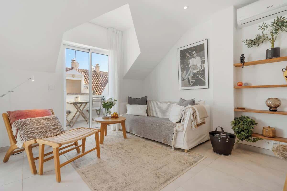 Ein modernes Wohnzimmer mit Sofa, einer Holzbank, einem kleinen Tisch und einer Stehlampe. Ein großes Fenster bietet einen Blick nach draußen.