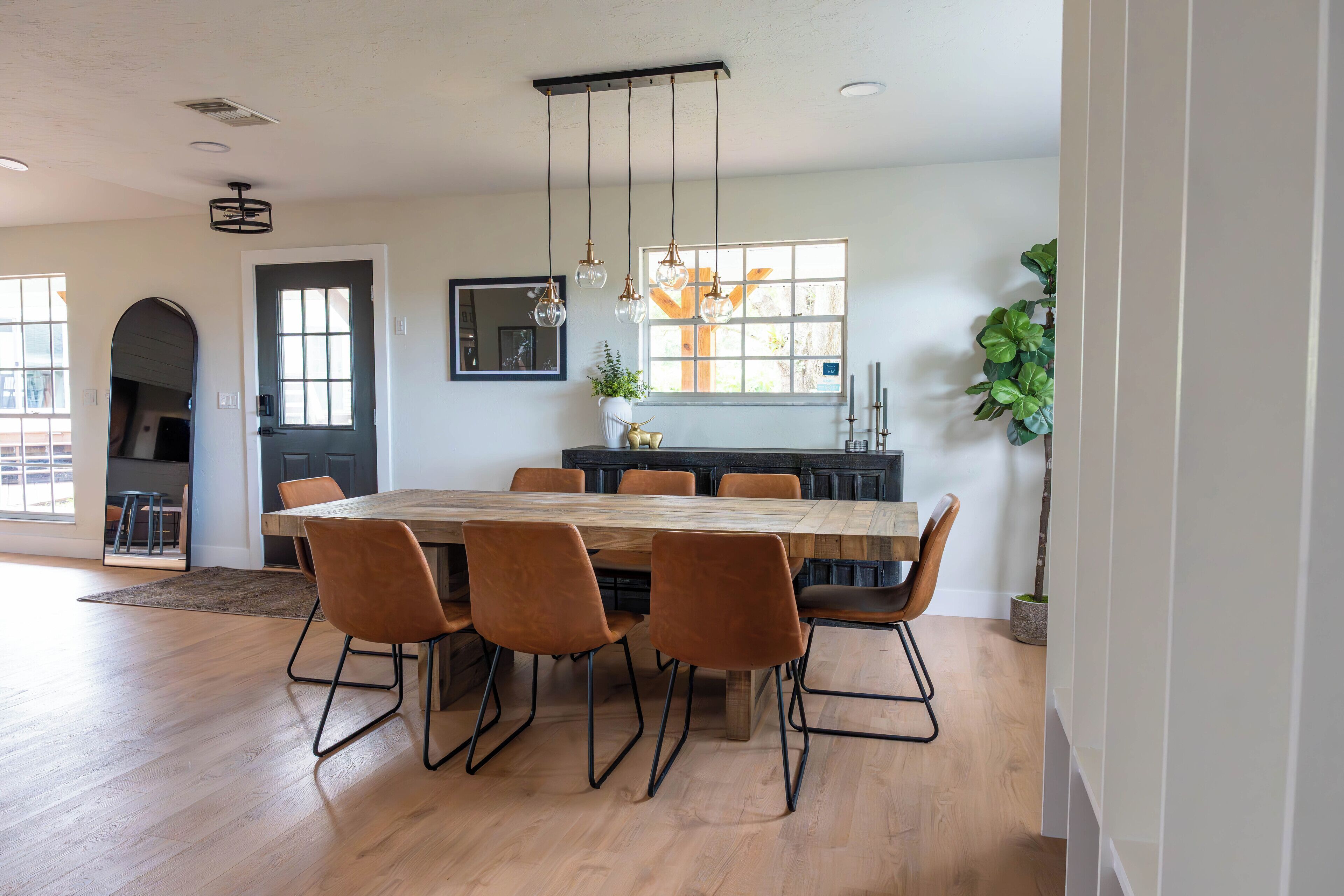 A spacious dining area with a large wooden table and six chairs, a modern pendant light, and a view of the outdoors through a window.