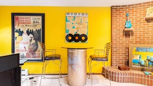 A room with a yellow wall, a poster of Moulin Rouge, a piano, two chairs, a table, and a brick wall with a guitar.