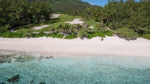 Una playa con agua turquesa cristalina, arena blanca y un campo de golf rodeado de vegetación exuberante y una montaña al fondo.