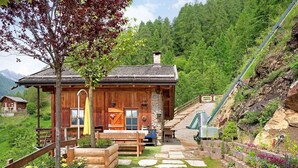 Une cabane en bois avec des marches en pierre menant à une terrasse en bois, entourée d’une végétation luxuriante et avec une chaîne de montagnes en toile de fond.