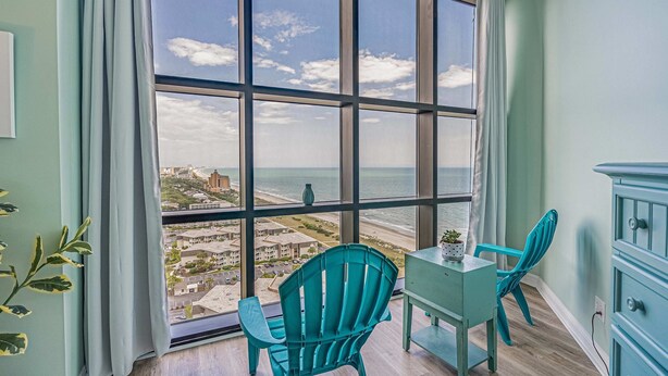 Ein Zimmer mit großem Fenster und Blick auf Strand und Stadt. Zwei türkisfarbene Stühle und ein kleiner Tisch sind vorhanden.