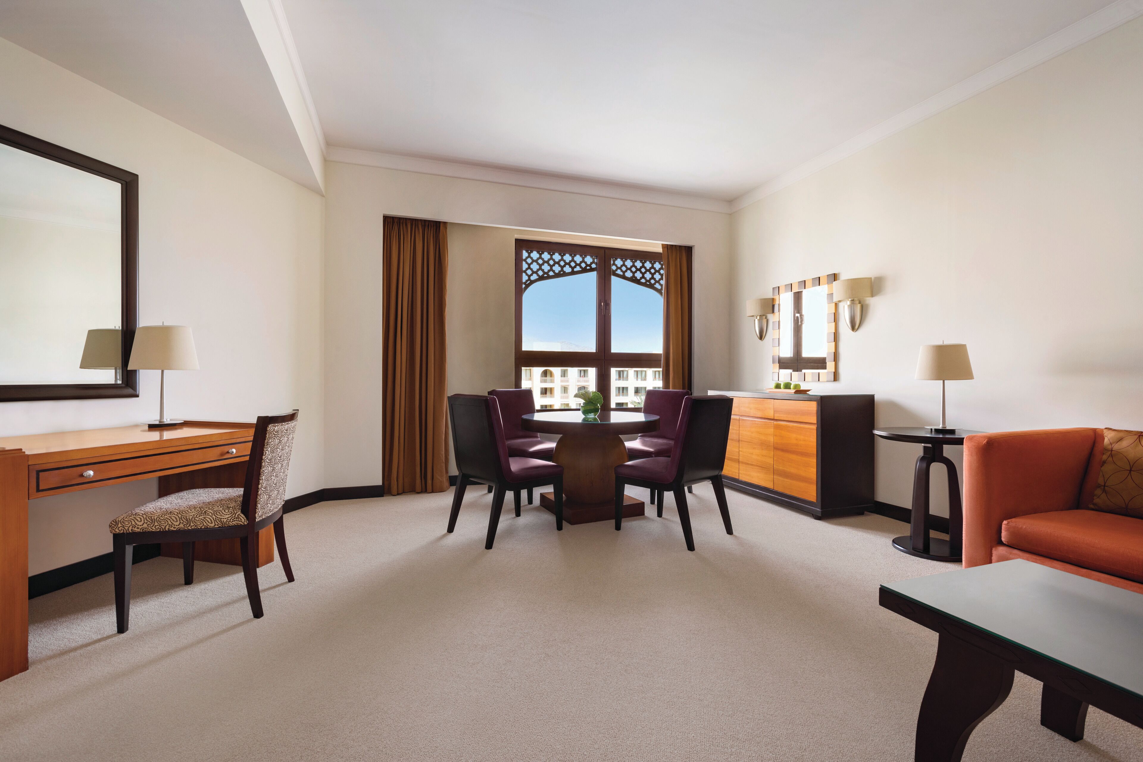A hotel room with a desk, two chairs, a round table, a sofa, a dresser, and a large window with a view.