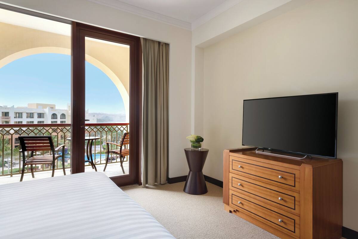 A hotel room with a bed, a TV on a wooden dresser, a balcony with chairs and a table, and a view of buildings.