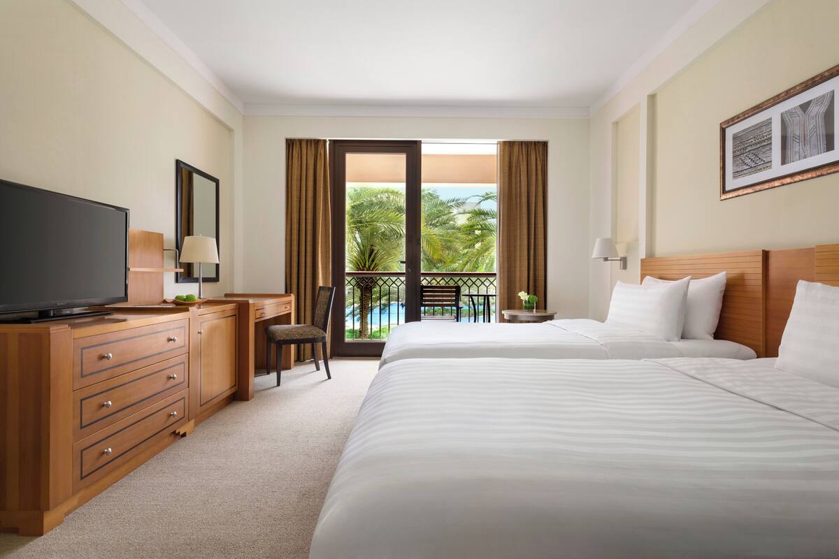 A hotel room with a large bed, a TV, a desk with a lamp, and a balcony with a view of palm trees.