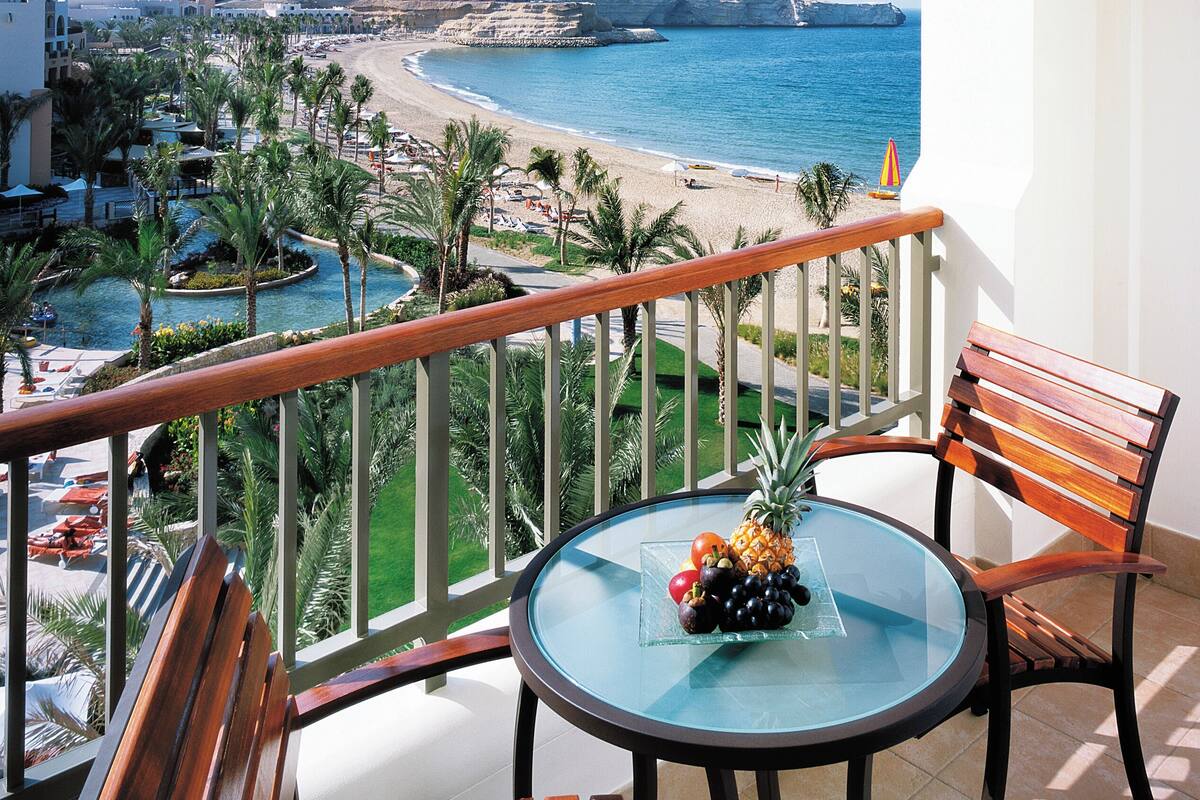 A balcony with a glass table, wooden chairs, and a view of the beach and ocean.