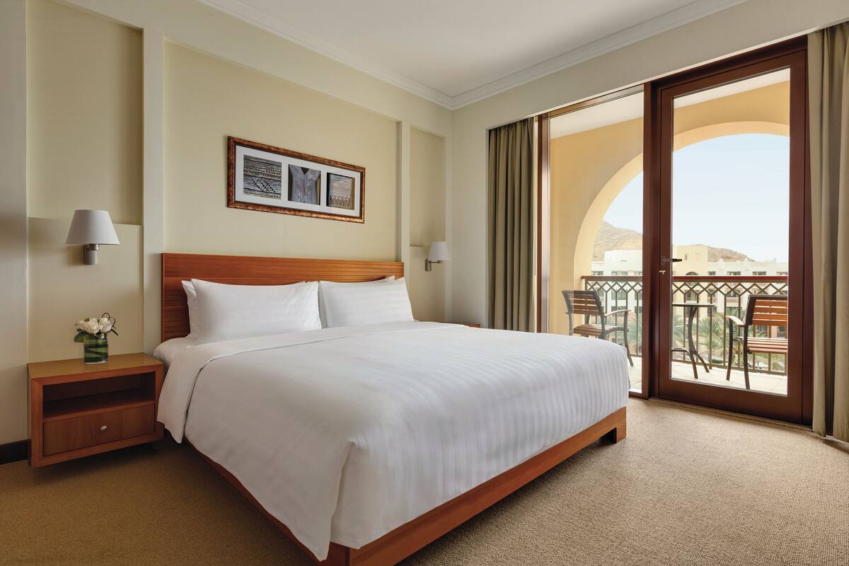 A hotel room with a large bed, a nightstand with a lamp, a framed picture on the wall, and a balcony with outdoor furniture.