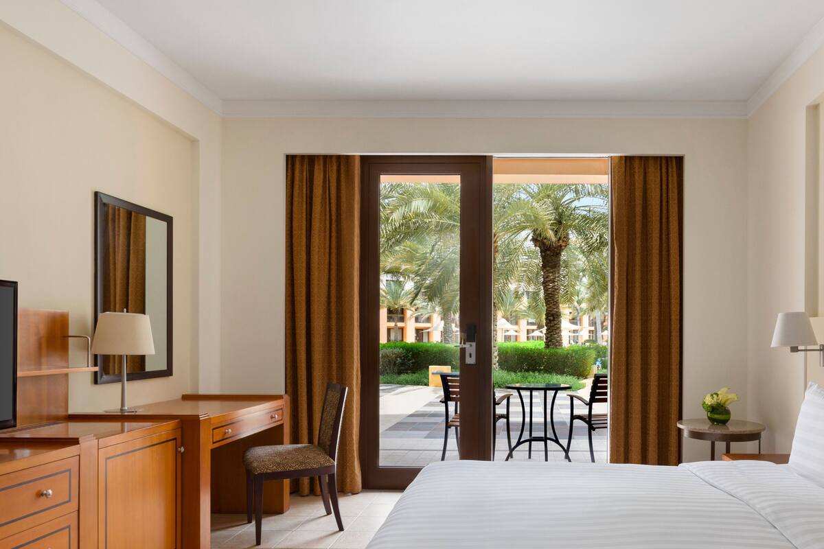A hotel room with a bed, a desk, a chair, a TV, and a view of palm trees and a pool outside.
