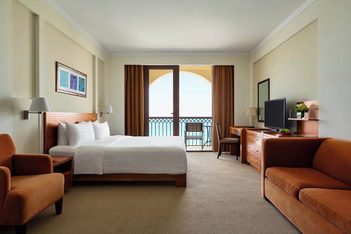 A hotel room with a bed, two armchairs, a desk with a television, and a balcony with curtains.