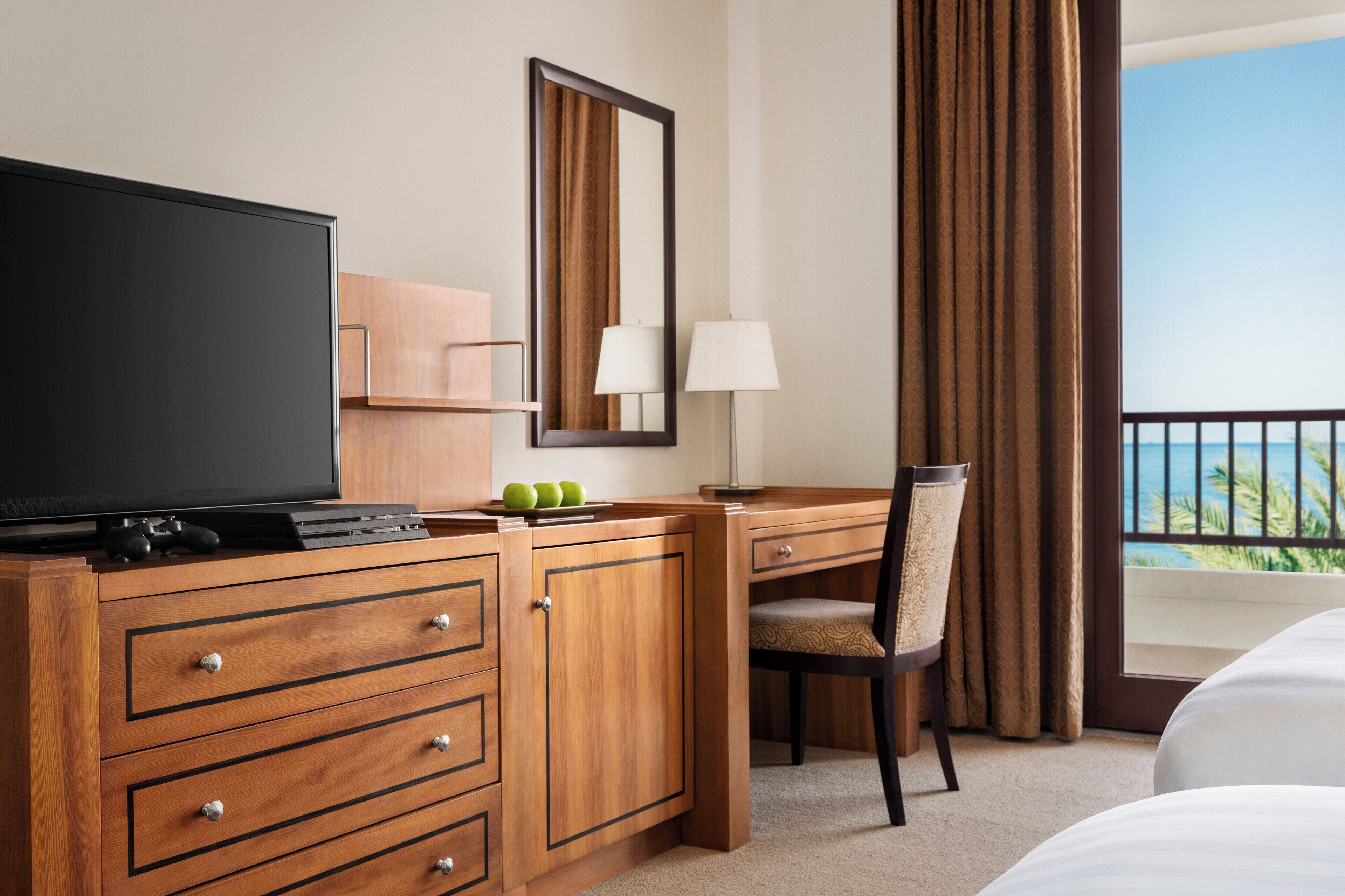 A hotel room with a wooden desk, a television, a chair, a bed, a window with curtains, and a view of the ocean.