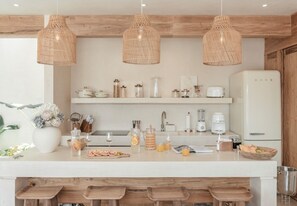 Eine moderne Küche mit weißen Arbeitsplatten, Holzbarhocker, hängenden Korbtreleuchtern und einem weißen Kühlschrank.