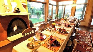 A dining room with a long table set for a meal, a fireplace, and large windows offering a view of the outdoors. - A touch of the wild west and the loving touch of the pioneer  (Bigfork)