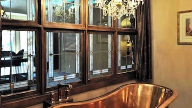 Quarto com uma banheira de cobre grande e em formato oval, grandes janelas com molduras escuras e um lustre.