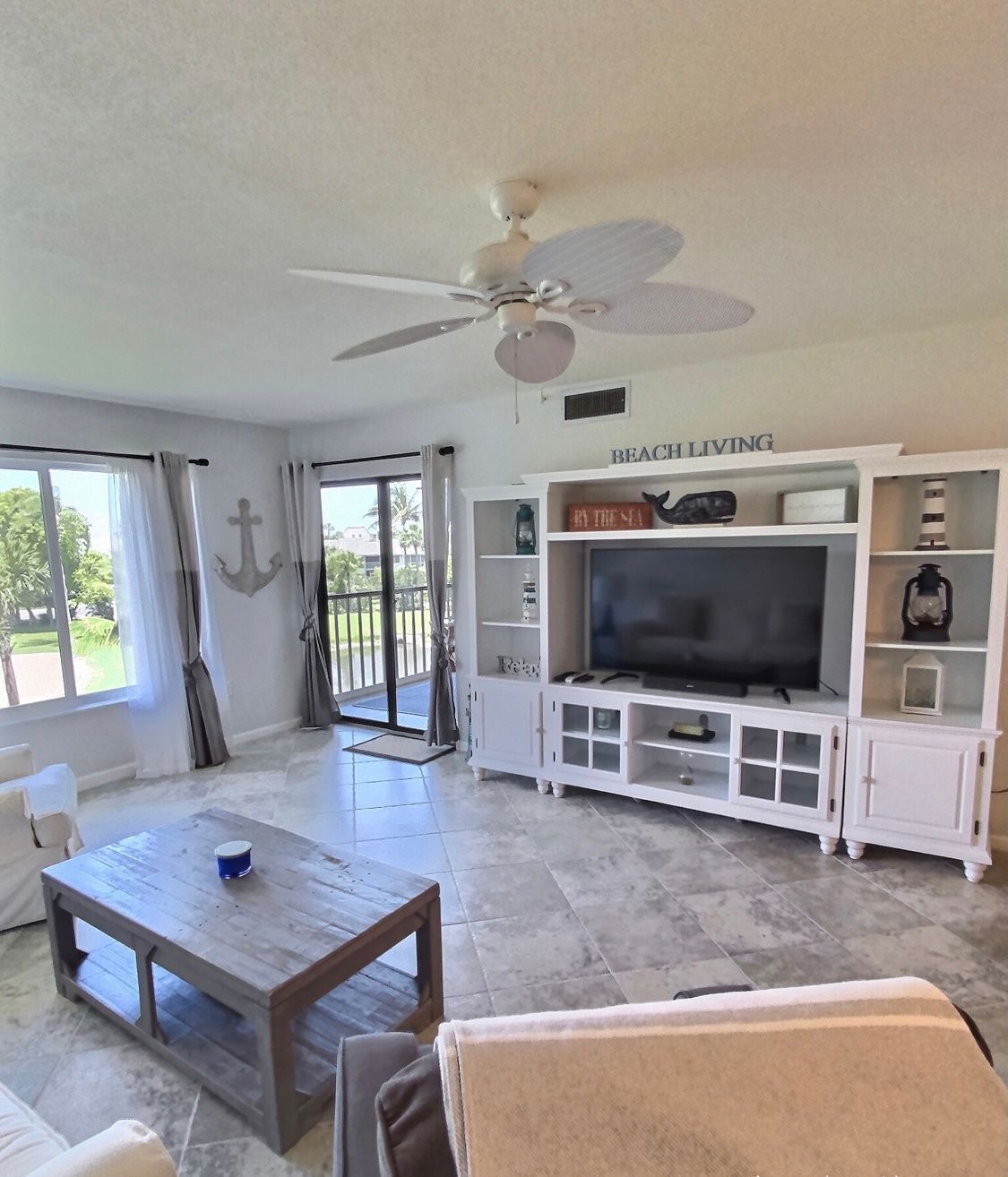 A spacious living room with a large flat-screen TV, a ceiling fan, and a sliding glass door leading to a balcony.