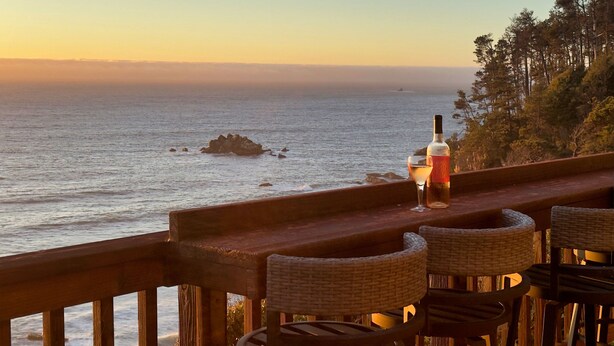 Un bar frente al mar con sillas de mimbre y una botella de vino sobre una barandilla de madera que da al océano.