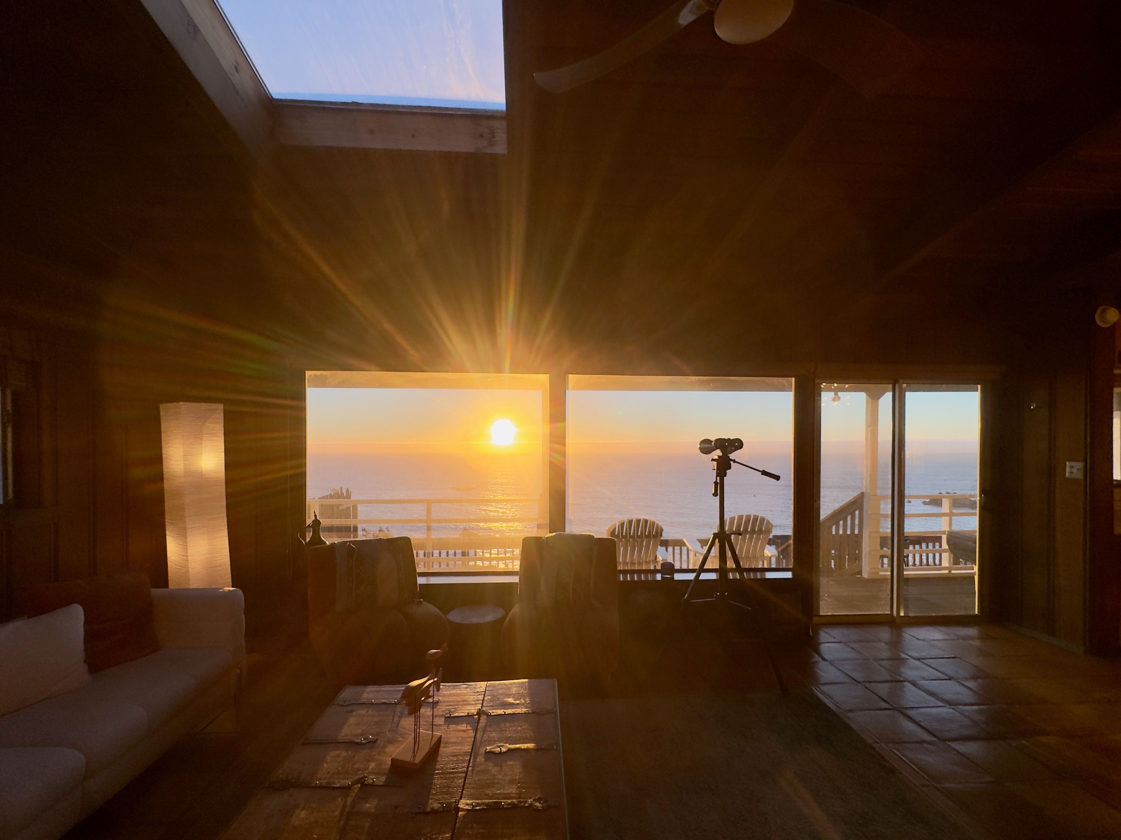 A room with a large window overlooking a sunset, a tripod with a camera, and a cozy seating area with a sofa and chairs.