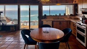 Una cocina con vista al océano, armarios de madera, una mesa redonda con sillas y una lámpara moderna.