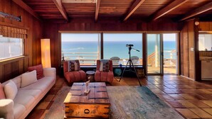 Una habitación acogedora con techo de madera, ventilador de techo, un amplio ventanal con vista al océano, un sofá, una mesa de centro y un trípode con una cámara.
