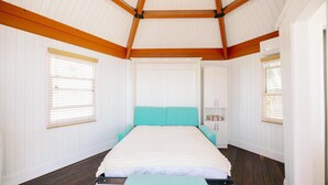 Une chambre à coucher avec un lit, des poutres en bois, des murs blancs et une fenêtre avec des stores.