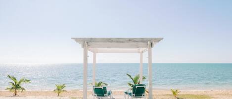 A beach with a white gazebo, two green chairs, and a small table.