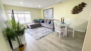 Een moderne woonkamer met een grijze modulaire bank, een eettafel met witte stoelen, een blauw-wit tapijt en een grote potplant.