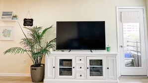 Een woonkamer met een flatscreen televisie op een wit kastje, een potplant en een deur met uitzicht naar buiten.