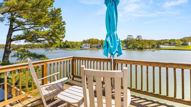 A wooden deck with two white chairs and a blue umbrella, overlooking a calm lake with houses in the background.
