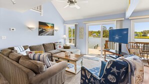 A living room with a sofa, coffee table, and a TV. There is a balcony with a view of the ocean and a sign that says 'LEWES OCEAN SIDE BAYSIDE.'