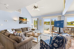 A living room with a sofa, coffee table, and a TV. There is a balcony with a view of the ocean and a sign that says 'LEWES OCEAN SIDE BAYSIDE.'